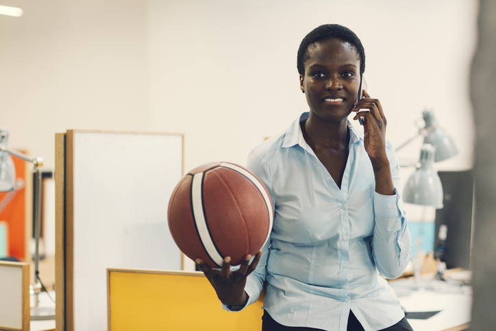 Smiling African businesswoman sitting on desk with basketball ball and talking on the phone in modern office. Front view, selective focus to her smiling face.